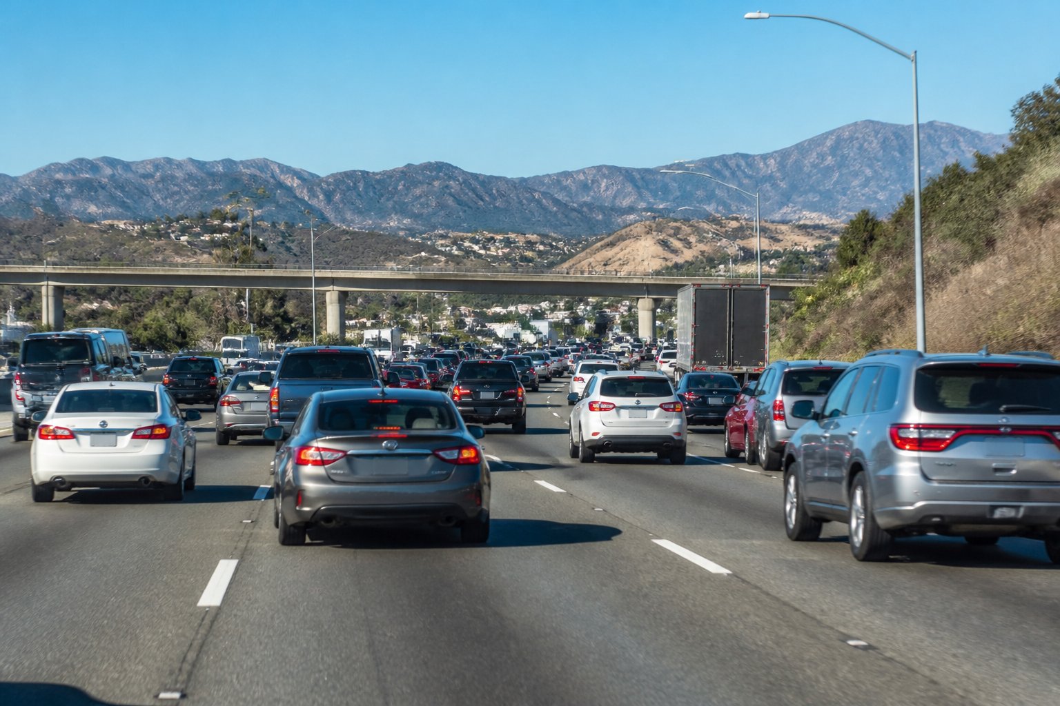 A busy multi-lane freeway with cars and trucks driving closely together, some changing lanes and braking, surrounded by urban infrastructure and hills in the background.